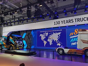 Gro&szlig;e Messeb&uuml;hne mit zwei Mercedes-Benz Lkws. Links ein bunt beklebter Lkw auf einem Podest, rechts das Heck einer wei&szlig;-orangen Zugmaschine. Im Hintergrund leuchtet eine blaue LED-Wand mit einer Weltkarte unter dem Banner 130 YEARS TRUCKS.