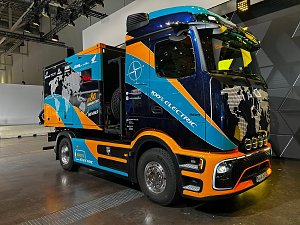 Blau-orangefarbener Mercedes-Benz Lkw schr&auml;g von vorne auf einem gl&auml;nzenden Hallenboden. Auf der Front ist eine Weltkarte aufgedruckt, auf der Seite steht der wei&szlig;e Schriftzug 100% ELECTRIC. Hinten ist ein bunter Kofferaufbau montiert.