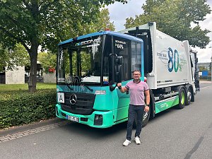 Ein Mann steht l&auml;chelnd neben einem modernen, hellgr&uuml;n-blauen Mercedes-Benz M&uuml;llwagen. Der wei&szlig;e Lkw-Aufbau hinten tr&auml;gt die gro&szlig;e Aufschrift 80%. Das Fahrzeug steht am Stra&szlig;enrand vor gr&uuml;nen B&auml;umen und einer Hecke.