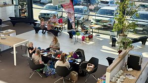 Besucherinnen sitzen an einem Tisch im Autohaus und beschäftigen sich mit bunten Häkelarbeiten. Im Hintergrund basteln Kinder.
