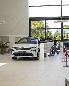 Ein weißer Škoda Enyaq Coupé steht im hellen Showroom der Autowelt Peter. Im Hintergrund betrachten Besucher weitere Fahrzeuge.