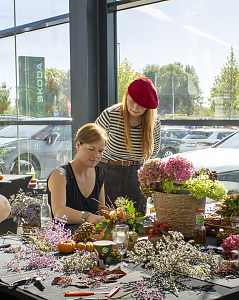 Zwei Frauen gestalten an einem Tisch herbstliche Blumenarrangements aus Hortensien, Zweigen, Beeren und kleinen Kürbissen.