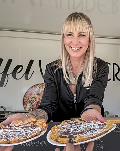 Lächelnde Mitarbeiterin eines Gastronomiestands reicht zwei frisch gebackene Waffeln mit Schokolade und Puderzucker über die Theke.