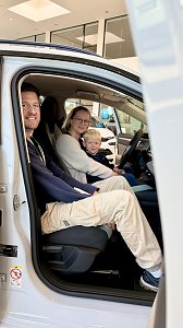 Familie mit Kind sitzt gemeinsam in einem weißen Škoda und testet den Innenraum des Fahrzeugs im Autohaus.