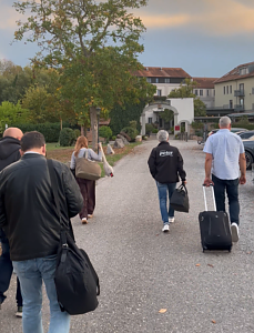 Mehrere Personen gehen von hinten zu sehen mit Gep&auml;ck einen asphaltierten Weg entlang. Links l&auml;uft eine Frau mit Schultertasche, rechts ein Mann in schwarzer Jacke mit Peter-Schriftzug sowie ein Mann, der einen Rollkoffer zieht. Im Hintergrund steh
