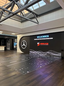 Eine gro&szlig;e, beleuchtete Medienwand steht in einer modernen Halle mit einer verglasten Deckenkonstruktion und dunklem Holzboden. Auf dem Bildschirm prangt das Mercedes-Benz-Logo, der Schriftzug Willkommen und das Logo von Ehrlich Logistics.
