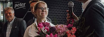 Lilly Hecker, frisch ausgebildete Automobilkauffrau und Jahrgangsbeste, steht lächelnd mit einem Blumenstrauß neben Geschäftsführer Andreas Peter, der in ein Mikrofon spricht. Im Hintergrund sind weitere Personen und das Logo der Autohaus-Peter-Gruppe zu sehen. (Autohaus Peter)