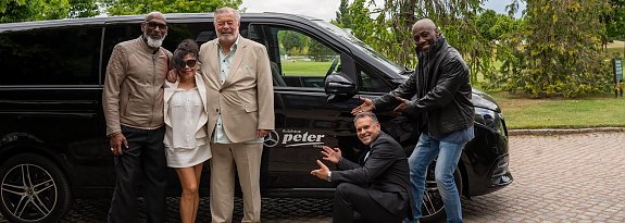 Gruppenfoto vor Shuttlebus: F&uuml;nf Personen, darunter vier M&auml;nner und eine Frau, posieren lachend vor einem schwarzen Mercedes-Benz Shuttlebus mit dem Logo Autohaus Peter.