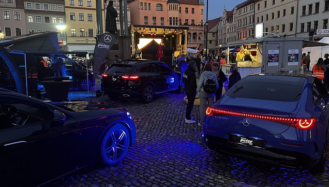 Das Bild zeigt die Eindrücke der Aftershowparty. Die beleuchteten Mercedes-Benz Modelle strahlen in der Abenddämmerung mit Blick auf die Bühne der Veranstaltung. Zudem sind mehrere Interessierte Gäste an unseren Fahrzeugen im Gespräch.  