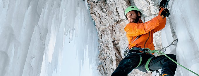 Mann mit orangefarbener Jacke und Helm klettert in vereisten Bergen.
