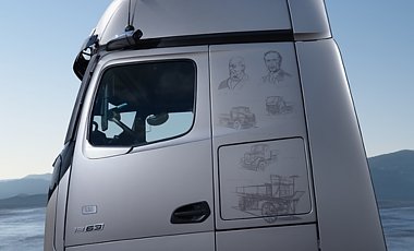 Silbernes Lkw-Fahrerhaus von der Seite vor blauem Himmel. Auf dem Lack hinter der Fensterscheibe sind feine Zeichnungen von historischen Autos und Portr&auml;ts von Carl Benz und Gottlieb Daimler.
