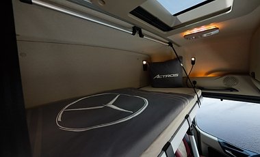Schlafkabine im Lkw-Fahrerhaus. Ein schmales Bett mit grauer Decke, auf der ein gro&szlig;er Mercedes-Stern prallt. Am Kopfende liegt ein graues Kissen mit dem Schriftzug Actros. Oben eine kleine Dachluke und warmes Licht aus einer Lampe.